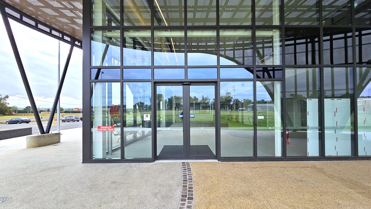 A concrete pavement adorns the concourse, with vehicle parking on the far left. In the middle stands a modern facility building of concrete and glass, featuring a large circular shade with steel mesh. The entrance boasts a floor-to-ceiling glass wall with an automatic sliding door. Chairs and bike racks are visible on the left side.