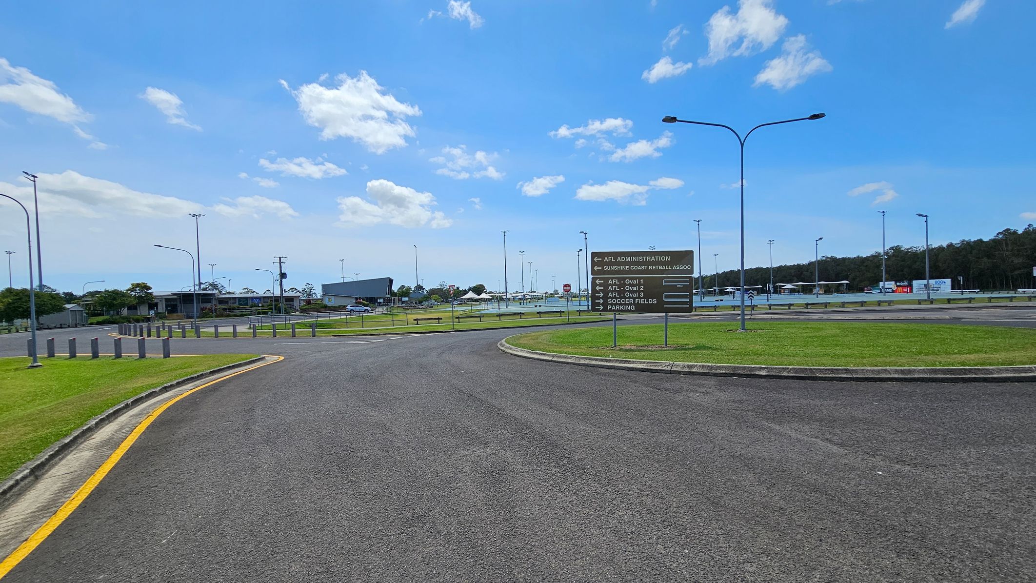 A paved road curves to the left, bordered by grass with a yellow line marking the edge. On the right, a sign lists directions to various sports facilities, including AFL ovals and soccer fields. In the background, there are several buildings, light poles, and a parking area. Trees line the horizon on the right side. The sky is partly cloudy.