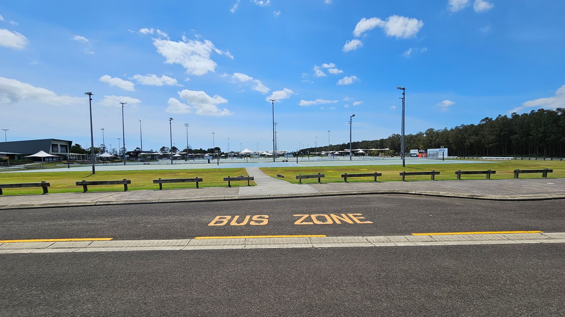 A paved road with "BUS ZONE" painted in yellow is in the foreground. Beyond the road, there is a grassy area with several wooden benches and a path leading to a sports complex. The complex features multiple light poles and a large building on the left. In the background, there are more structures and a line of trees on the right.