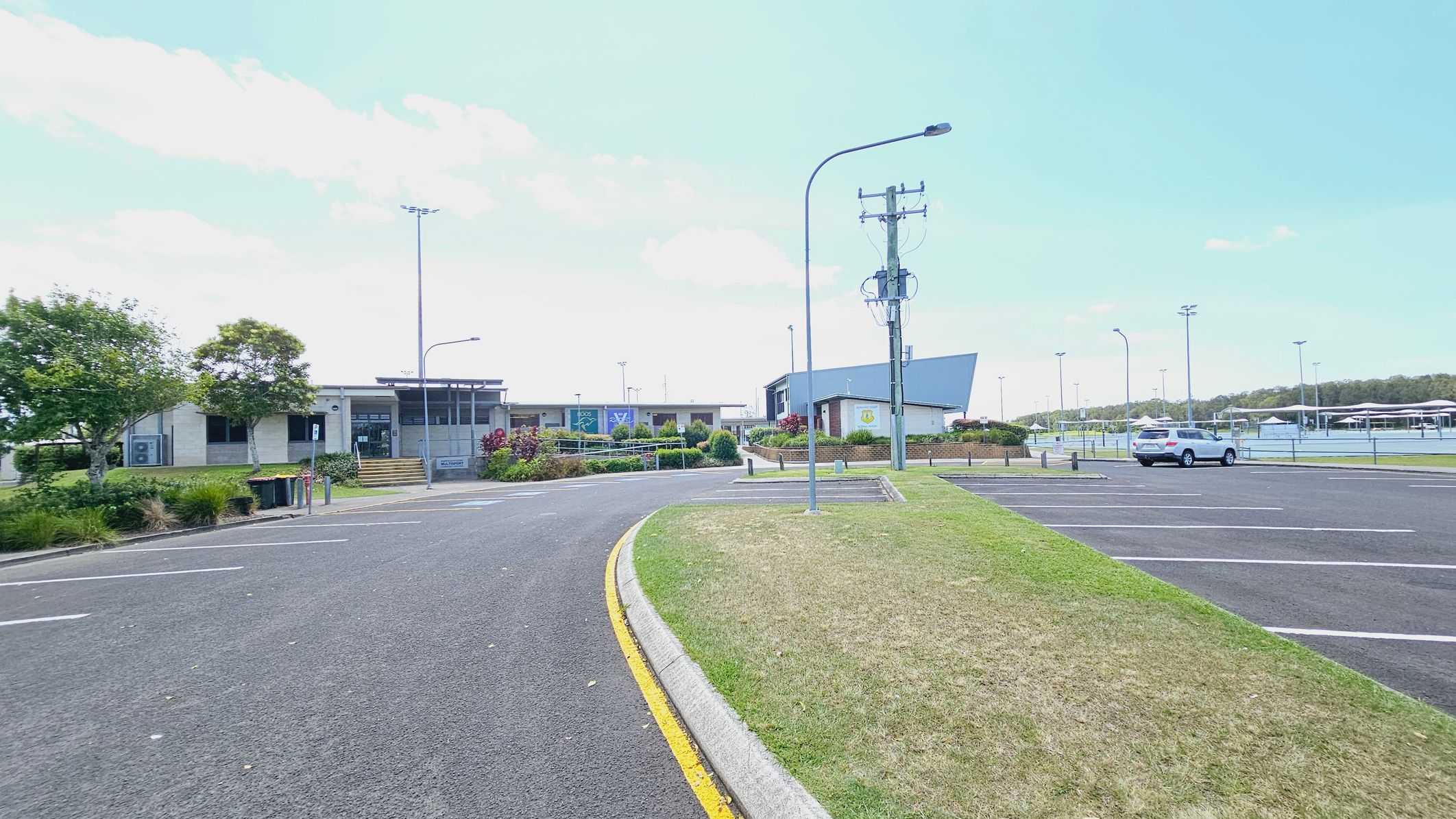 A car park with a single white vehicle on the right. The foreground features a curved road with a yellow line and adjacent grass. In the background, there are buildings with modern architecture, surrounded by landscaped greenery. Several tall streetlights and a utility pole are visible. Tennis courts with fences are on the far right.