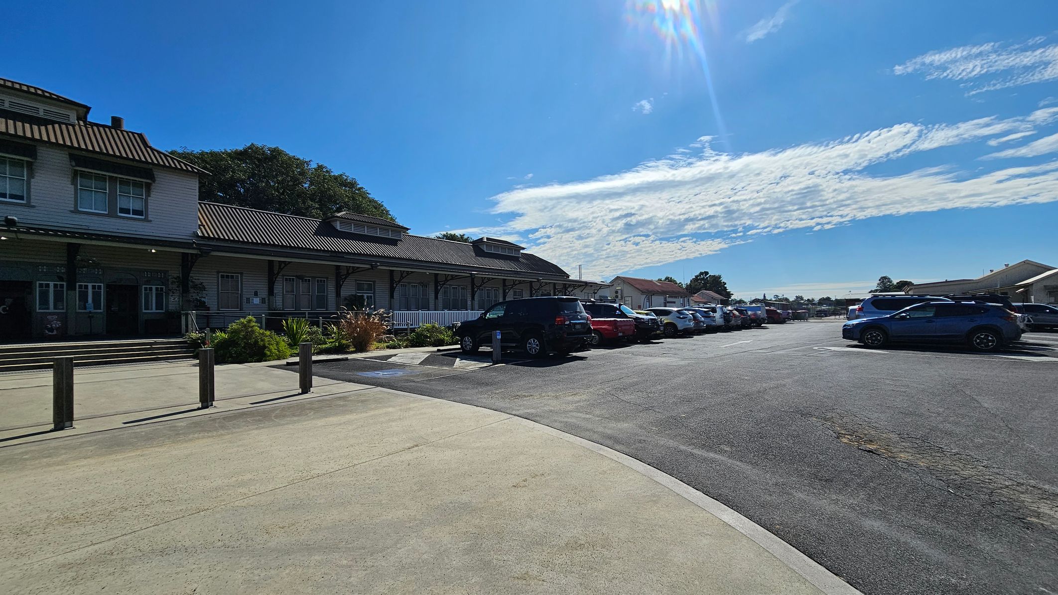 A paved area with a row of parked cars on the right, including a blue SUV in the foreground. On the left, a building with a dark metal roof and multiple windows, surrounded by some greenery. The building has a covered porch with steps leading up to it. The sky is clear with some clouds.