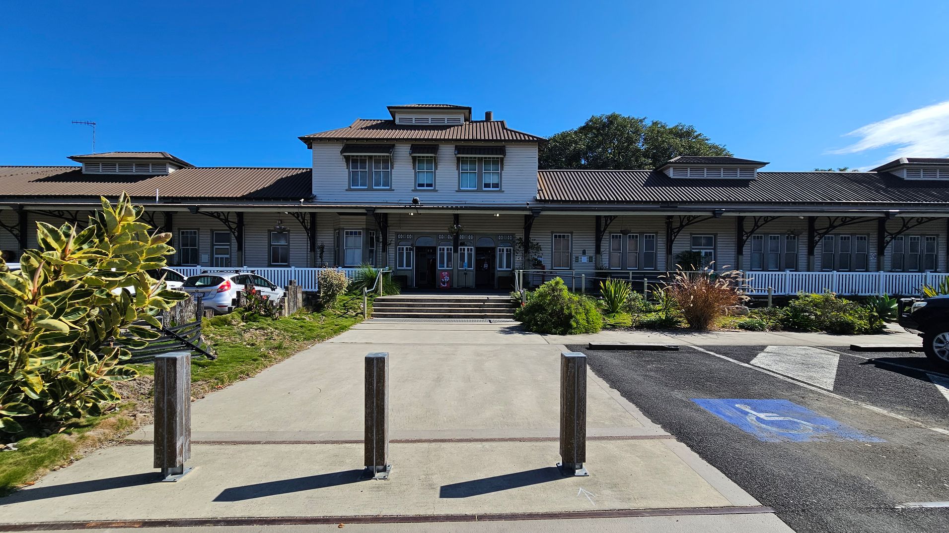 A large, two-storey building with a brown, corrugated roof and multiple windows. The entrance features a set of steps leading up to double doors. In front, there are three metal bollards on a paved path. To the left, a small garden with shrubs and a parked white car. On the right, a marked disabled parking space on the tarmac. The building is surrounded by greenery, including bushes and trees.