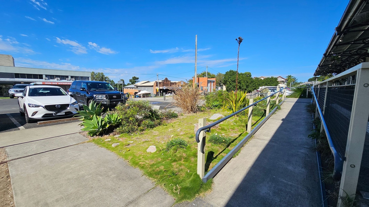 A concrete pathway with a metal railing runs along the right side, bordered by a grassy area with various plants and rocks. On the left, two parked cars, a white Mazda and a dark SUV, are visible. In the background, there are buildings with a mix of architectural styles, including a brick structure and a modern one. The sky is clear and blue.