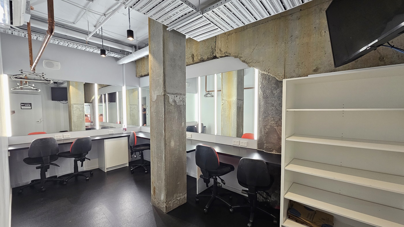 The dressing room features mirrors surrounding the walls. Adjustable chairs are positioned near the mirrors. To the right, there's a white display rack, with a flatscreen mounted on top. The ceiling displays exposed concrete beams, pipes, and air conditioning vents. The walls are white, and the floor is tiled black.