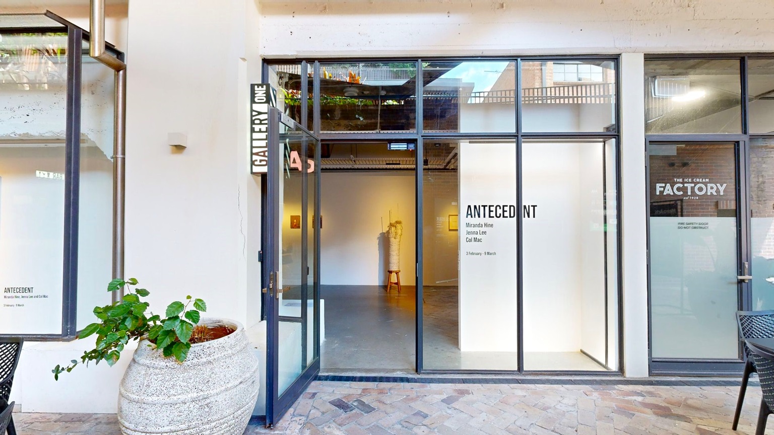 A manual glass door opens outward, with a signage reading "Gallery One" on the wall above. An ornamental plant sits in a huge pot below. Signages are affixed to the right side of the glass door. Movable black tables and chairs are arranged in front. The floor is tiled with brick, and the wall is white. Inside, a section of the gallery is visible.