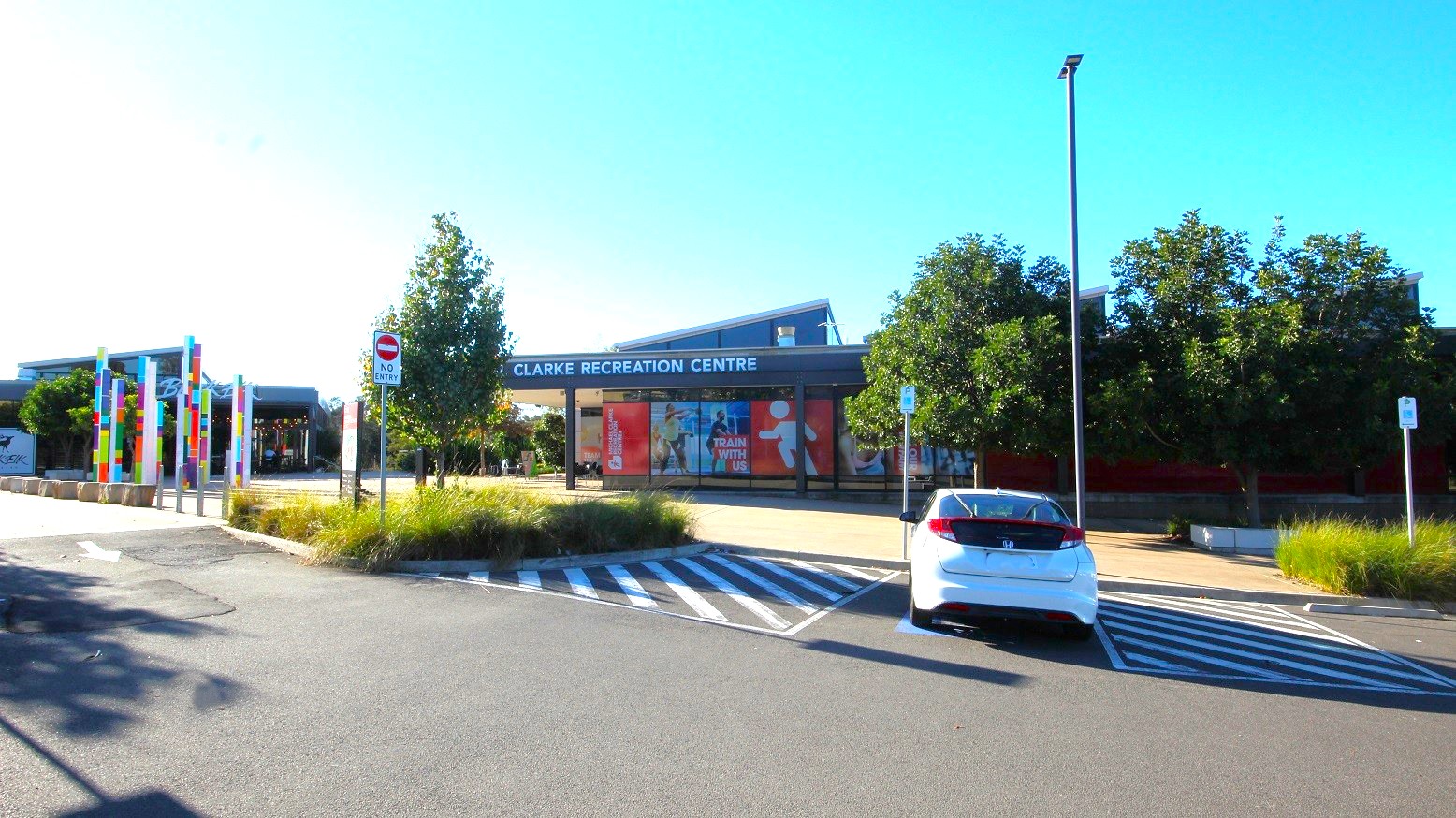 A white car is parked in one of the accessible parking lots in Michael Clarke Recreation Centre, marked by white lines and a blue accessible parking sign. A no-entry parking sign can be seen on the left side of the accessible parking area within the entry garden. Long but thin rectangular blocks with colorful designs are placed behind the sandstone blocks.