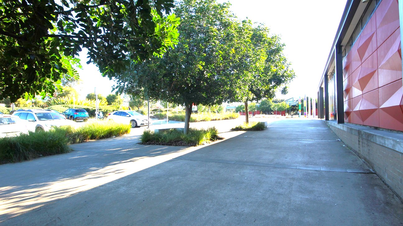 Step-free entry from one of the two accessible parking spaces at the Michael Clark Recreation Centre. The pathway is on the side of the building, painted in red, and has an elevated-looking diamond pattern. Surrounding the venue are medium-height trees with significant foliage planted within a square land area with parallel venation. Further on the left are a number of cars occupying the parking area.