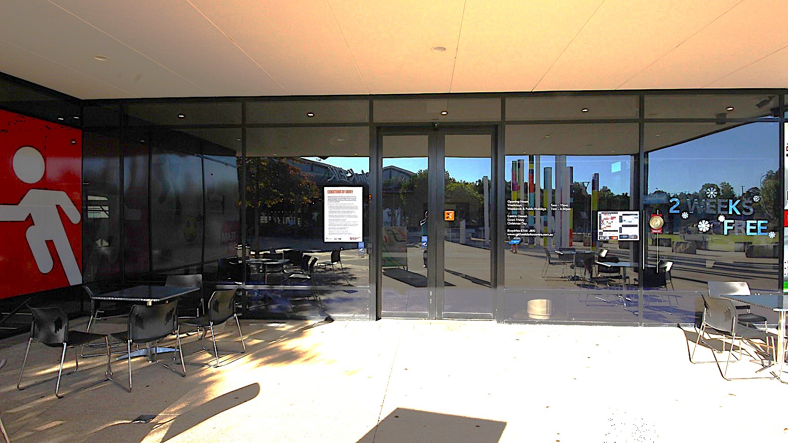 A closer look at the double glass door at Michael Clarke Recreation Centre. Each chair in the seating area close to the entrance is arranged surrounding a single table. On the left side of the door are the conditions of entry, while on the right are opening and closing days, the exact time the venue is open, and their contact information for any inquiries.