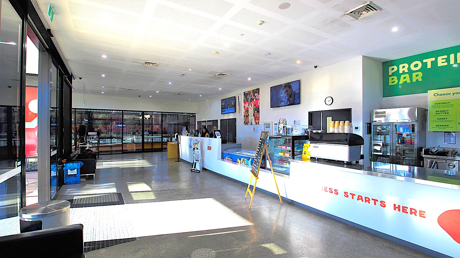 Overview of the entire reception area and Café as well the glass windows further to the left of the reception. The basketball center is visible through these glass windows. From this image, the double doors at the Michael Clarke Recreation Centre are automatic. There is a silver rubbish bin near the door, and several cold drinks stored in a large fridge which are available in the Café.