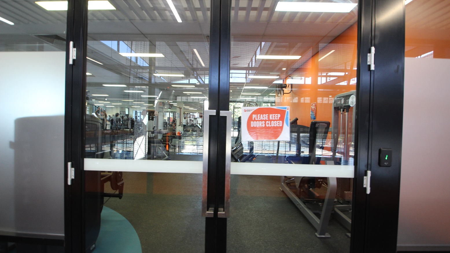 A “Please keep doors closed” orange with white and blue accents sign can be seen on the right side of the glass double door of the Gymnasium. People enjoying the different training equipment can be seen through the glass doors. At a glance, the distinct orange wall inside can be first noticed.