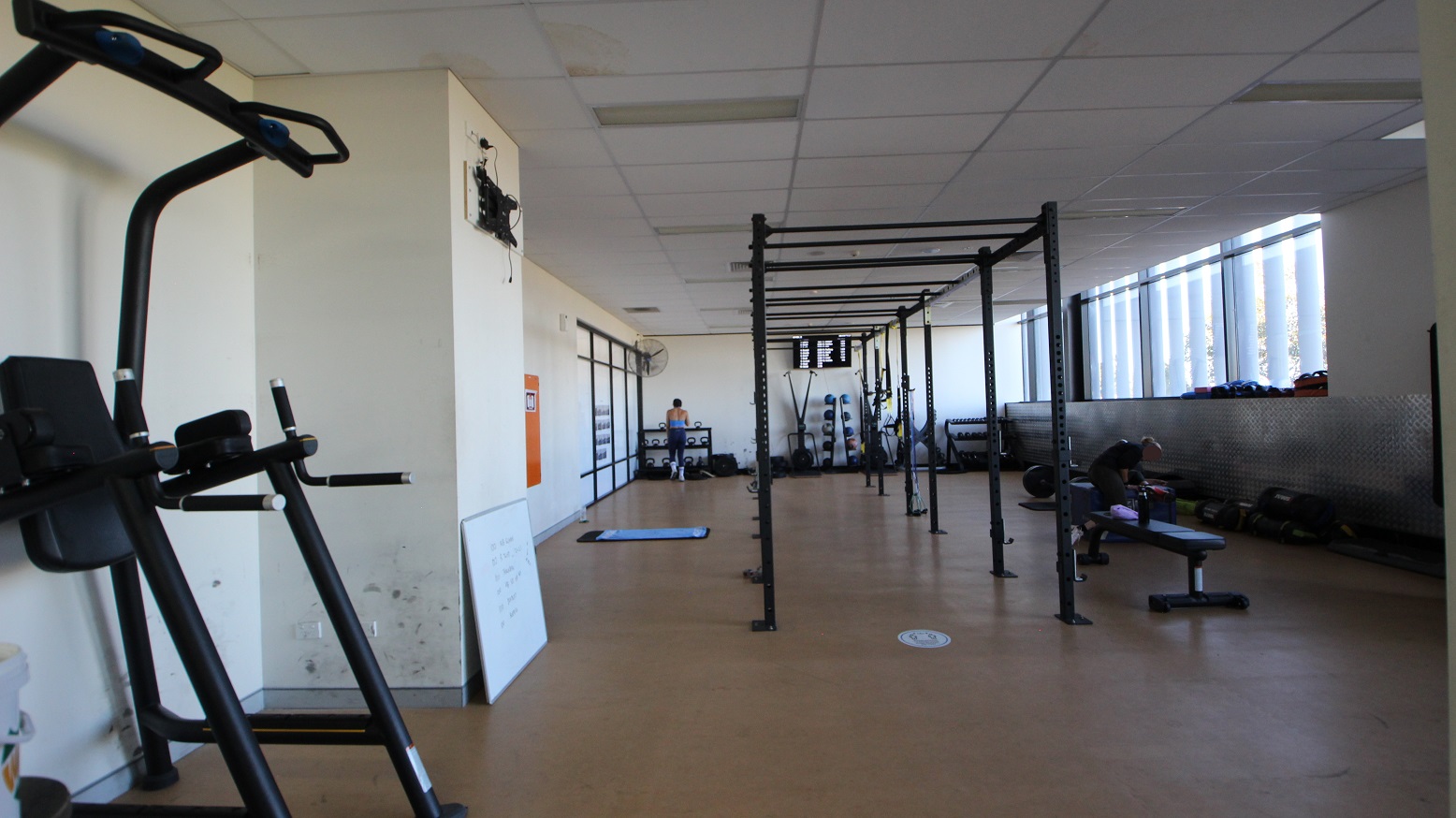 The image shows another part of the Gymnasium. The walls are painted white, and a shiny silver metal platform with a mid-waist height is placed against the right wall of the room. In the middle is a black crossfit rig and a bench. A medium size whiteboard is laid against the left wall, and different resistance training equipment is accessible further at the back.