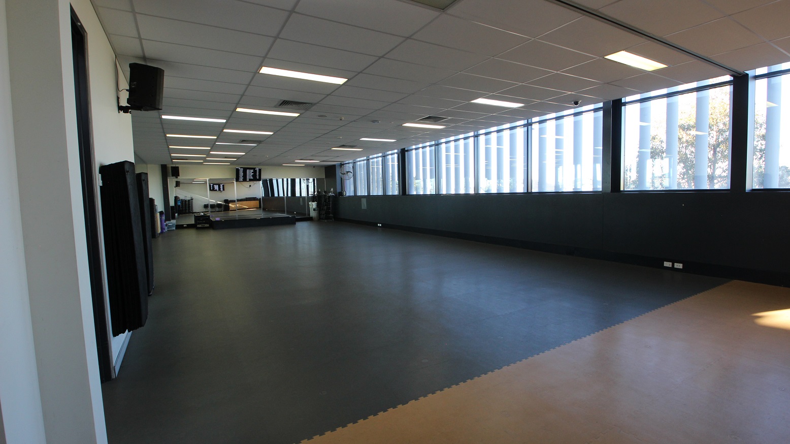 A room with a large open space covered with grey rubber mats in the Gymnasium of Michael Clarke Recreation Centre. Further to the back are floor-to-ceiling mirrors and a black elevated platform that looks like a small stage. On the left are speakers mounted on the wall.