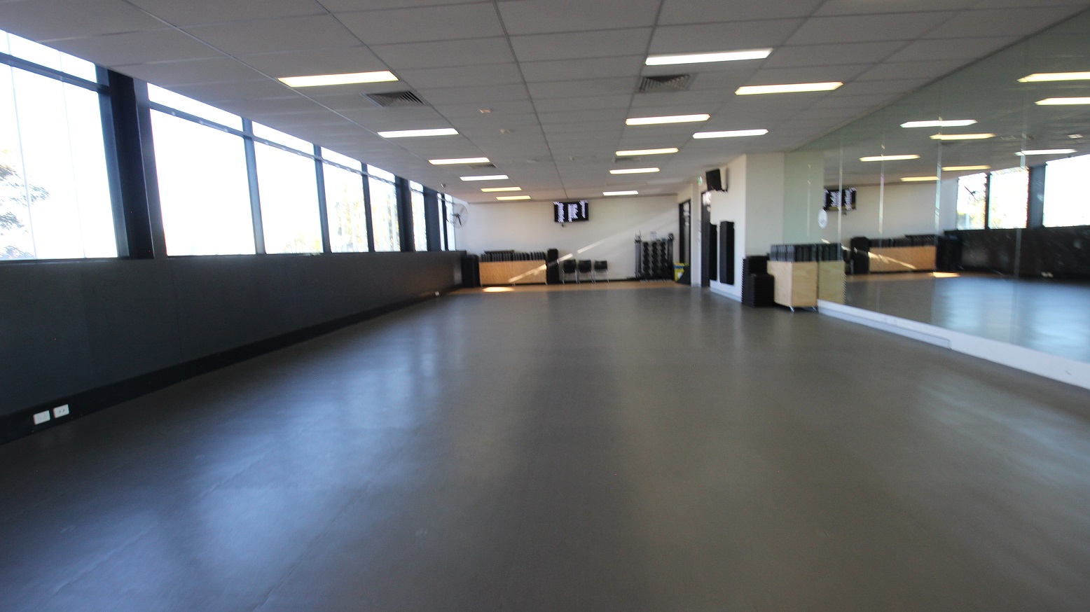 Opposite view of the previous image. This image shows that next to the wall with speakers are floor-to-ceiling mirrors covering this entire side of the wall. The ceiling is white, subdivided by square tile sections. The room is provided with a combination of natural lighting coming from the mid to upper glass windows and artificial lighting on the ceiling.