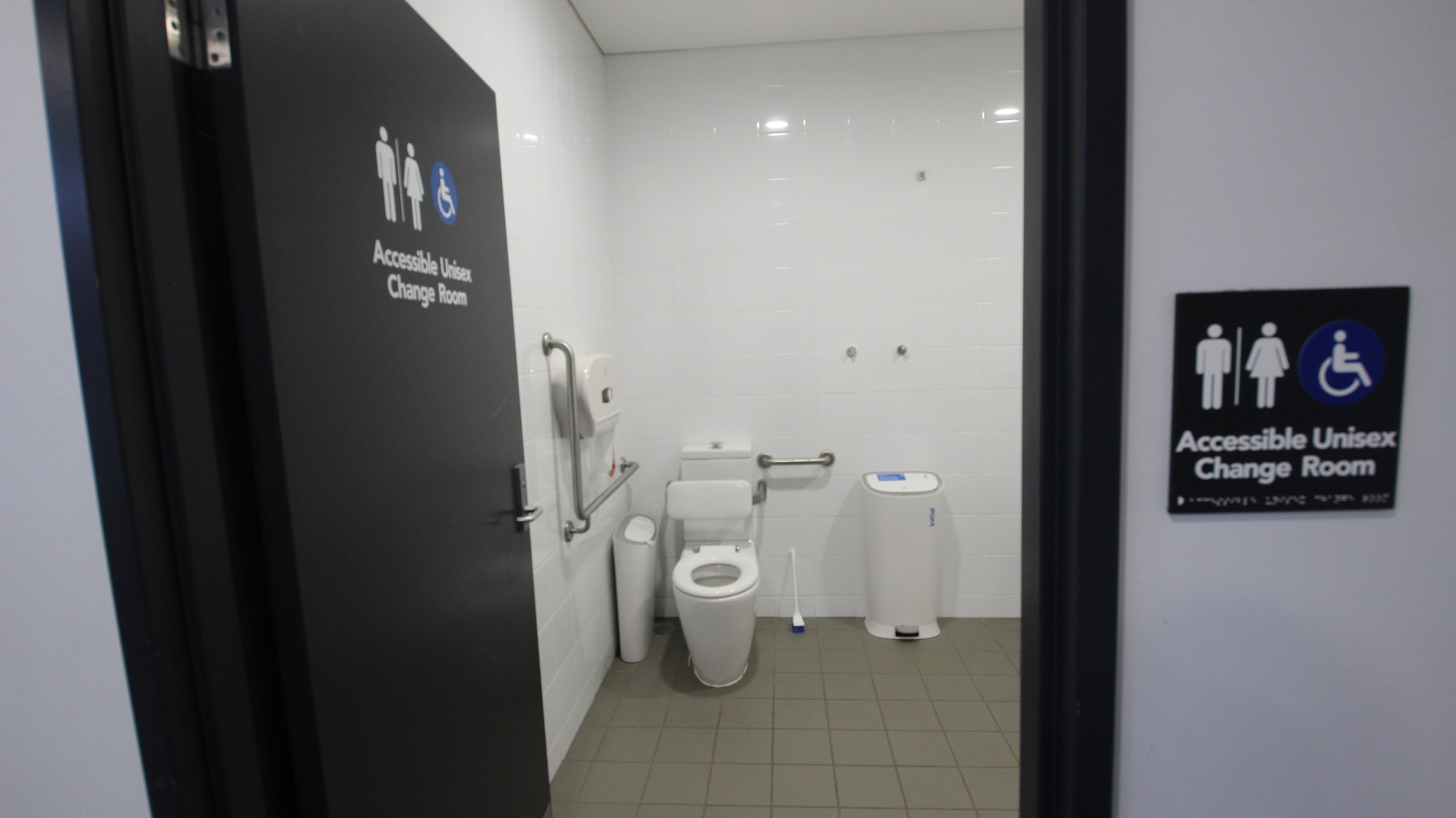 The image shows a part of the Accessible Unisex Change Room for persons with mobility disabilities in the Gymnasium. This change room can be distinguished by the blue accessible sign as well as male and female icons. The floor tiles are grey, while the wall tiles are white which makes the room polished and immaculate to the feeling. This image only shows the toilet bowl, white rubbish bin, toilet tissue paper holder, and blue toilet brush with white holder.