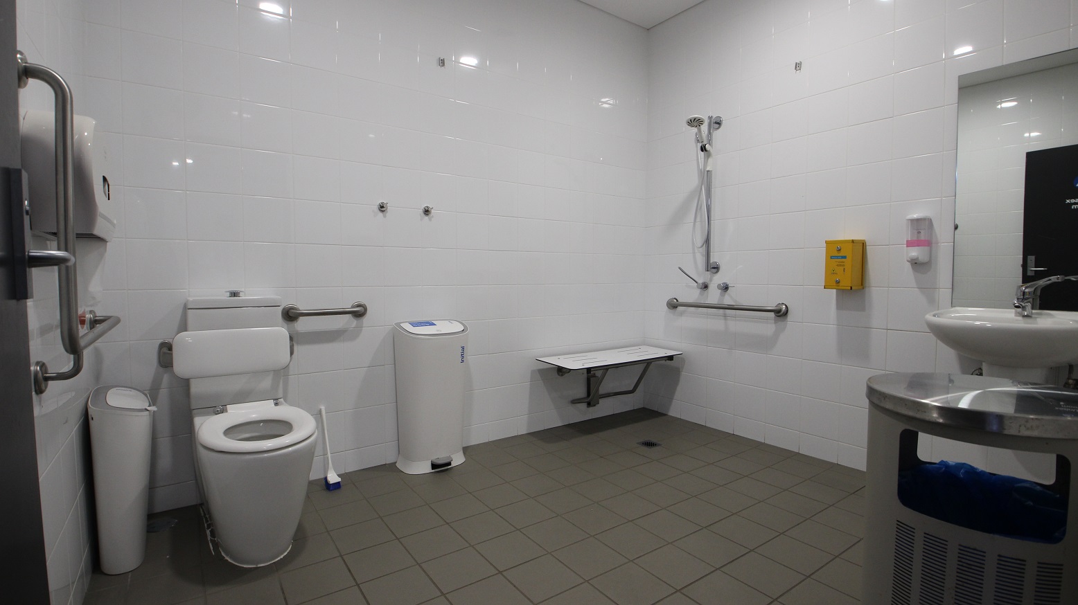 A complete view of the inside of the Accessible Unisex Change Room in the gymnasium. There is a white, hand-held shower, a white seating platform, and liquid soap dispenser mounted on the wall, a wash basin, and a metal rubbish bin.