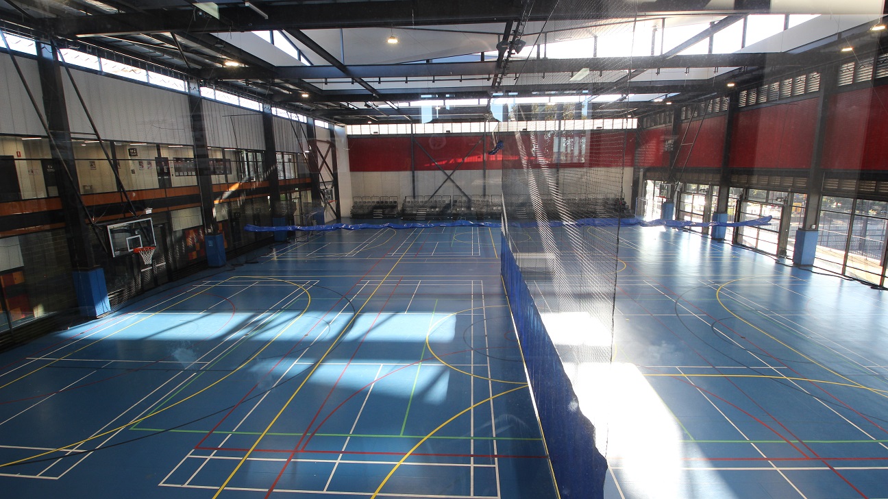 The upstairs offices and gymnasium overlook the stadium from above. The Stadium is the size of two basketball courts in a box shape warehouse with large, wide, steel black archways at the ceiling. The sloping roof and large skylights allow for significant natural lighting. The stadium has blue floors with white, yellow, black, and red lines. The space is separated by a black net with square holes and blue cloth at the bottom.