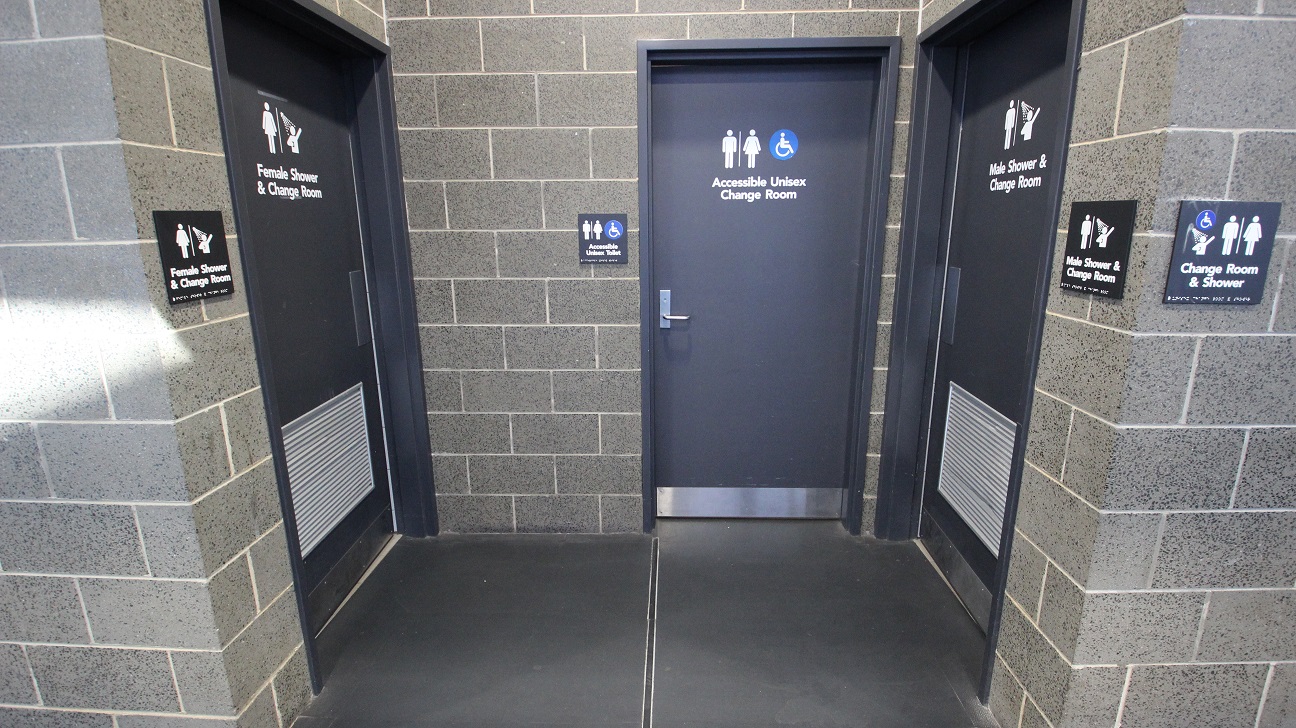 Accessible Unisex Change Room in the middle. It is different from the other changing room due to the blue and white accessible sign beside the male and female icons. On the opposite sides of the accessible unisex change room is the female shower and change room on the left and the male shower and change room on the right. Similar to the corridor walls of the previous image, this also has grey brick walls.