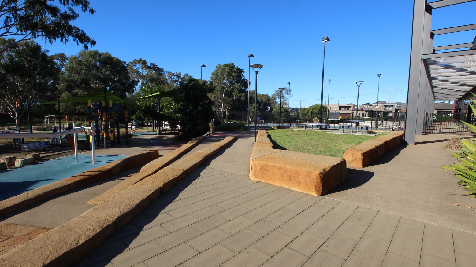 Tactile tiles with a slight elevation of brown concrete blocks on each side of the ramp. Several lamp posts, large trees with dense foliage, and benches can be seen throughout the area. A black tall fence surrounds the perimeter.