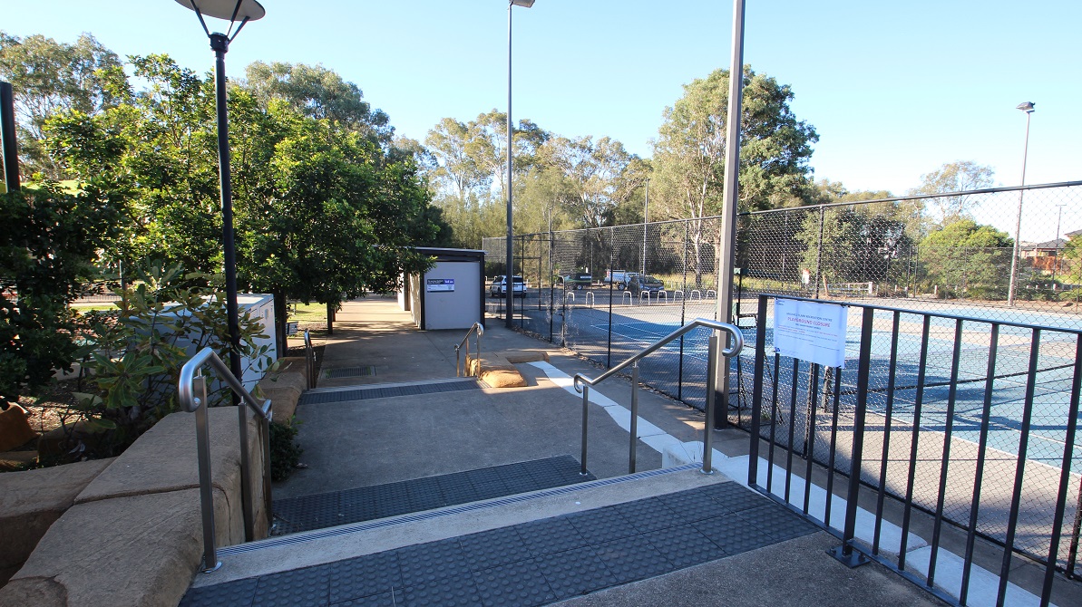 Staircase with handrails leading to the entrance of the Tennis Courts at the Michael Clarke Recreation Centre. The stairs are equipped with grey rubber mats that have circular elevations for better traction. The trees surrounding the area offer ample shade to the staircase