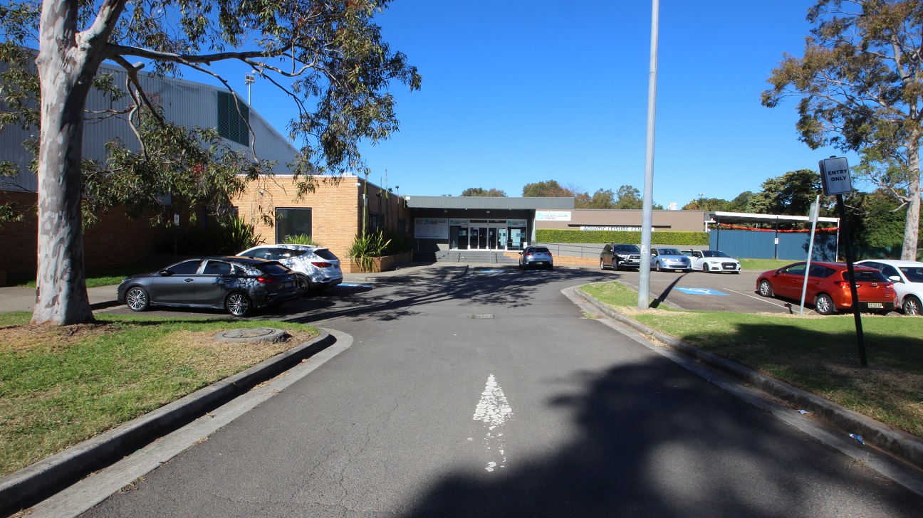The view metres away from the front of Michael Wendon Aquatic Leisure Centre. Michael Wenden ALC is a low-set brown and grey industrial, warehouse-style building. The accessible car parks available in front of the venue are marked by an accessible parking sign painted in white and blue. On the right side of the venue is a ramped access that allows for a step-free entrance with four grey steps to enter the venue through automatic glass sliding doors.