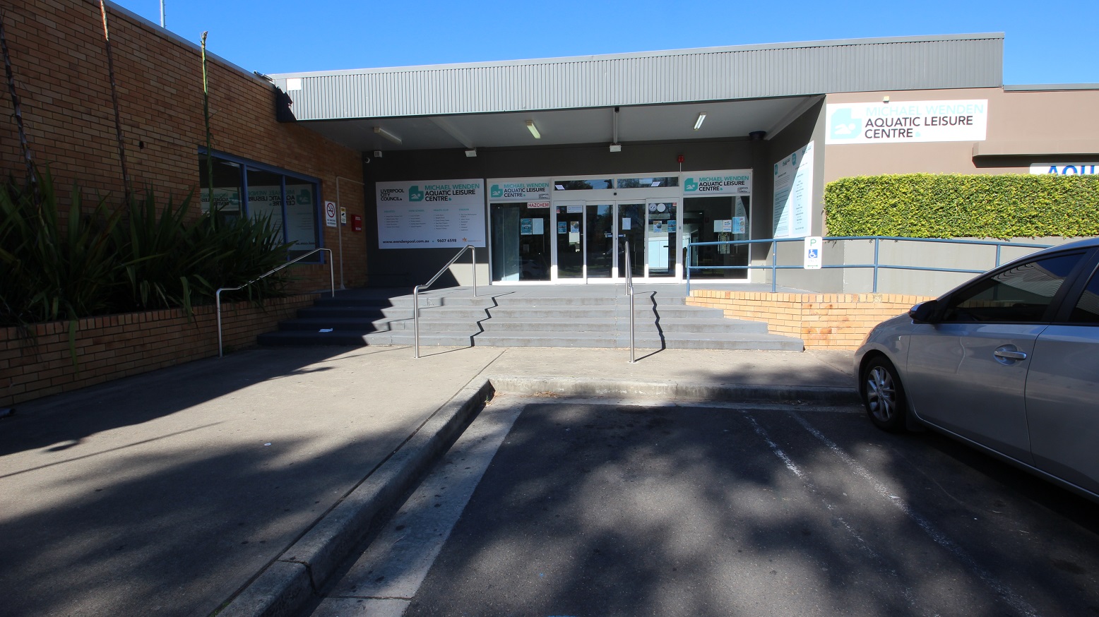 On the blue handrail near the automatic glass sliding doors is the accessible parking sign in white and blue. Bushes cleanly trimmed filled the garden against the smooth brown wall on the right side of the Michael Wendon Aquatic Leisure Centre. Several marketing and informative posters in different sizes are placed on the walls of the entrance doors. There is also a no-smoking sign, and a CCTV camera in operation can be seen on the brick wall to the left of the entrance wall.
