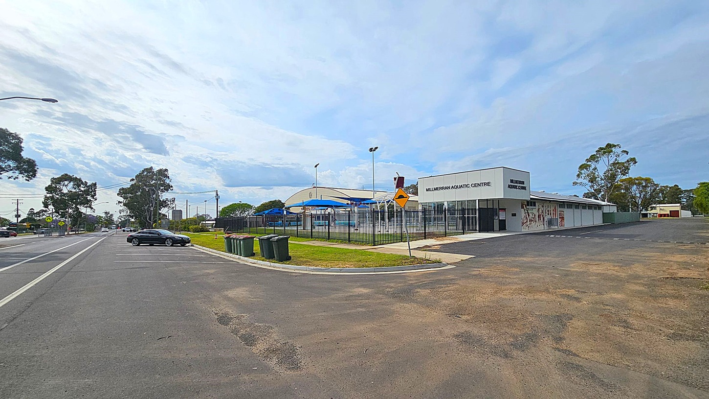 A street view with a parking area on the left, featuring a few parked cars and several rubbish bins. On the right, there's a building labelled "Millmerran Aquatic Centre" with a fenced pool area and blue shade structures. The background includes trees and a few more buildings. The ground is a mix of asphalt and grass, and the sky is overcast.