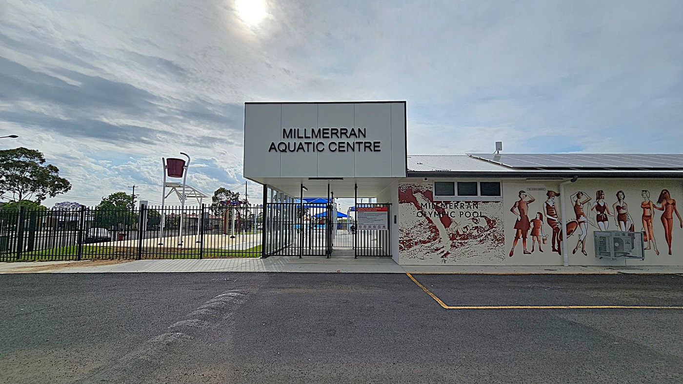 Entrance to the Millmerran Aquatic Centre with a large sign above the gate. To the right, a mural on the building wall depicts people in swimwear. The ground is paved with asphalt, and a black metal fence surrounds the area. In the background, a large bucket feature is visible, likely part of a water play area. Trees and a street are visible beyond the fence.
