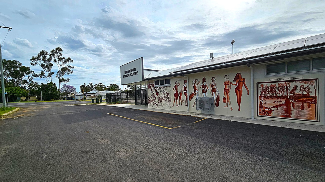 A building with a sign reading "Mulgildie Aquatic Centre" on the left side. The wall features a mural of people in vintage swimwear and a painting of a riverside scene. The foreground shows an empty asphalt parking area with yellow lines. In the background, there are trees and a few houses. The sky is overcast.