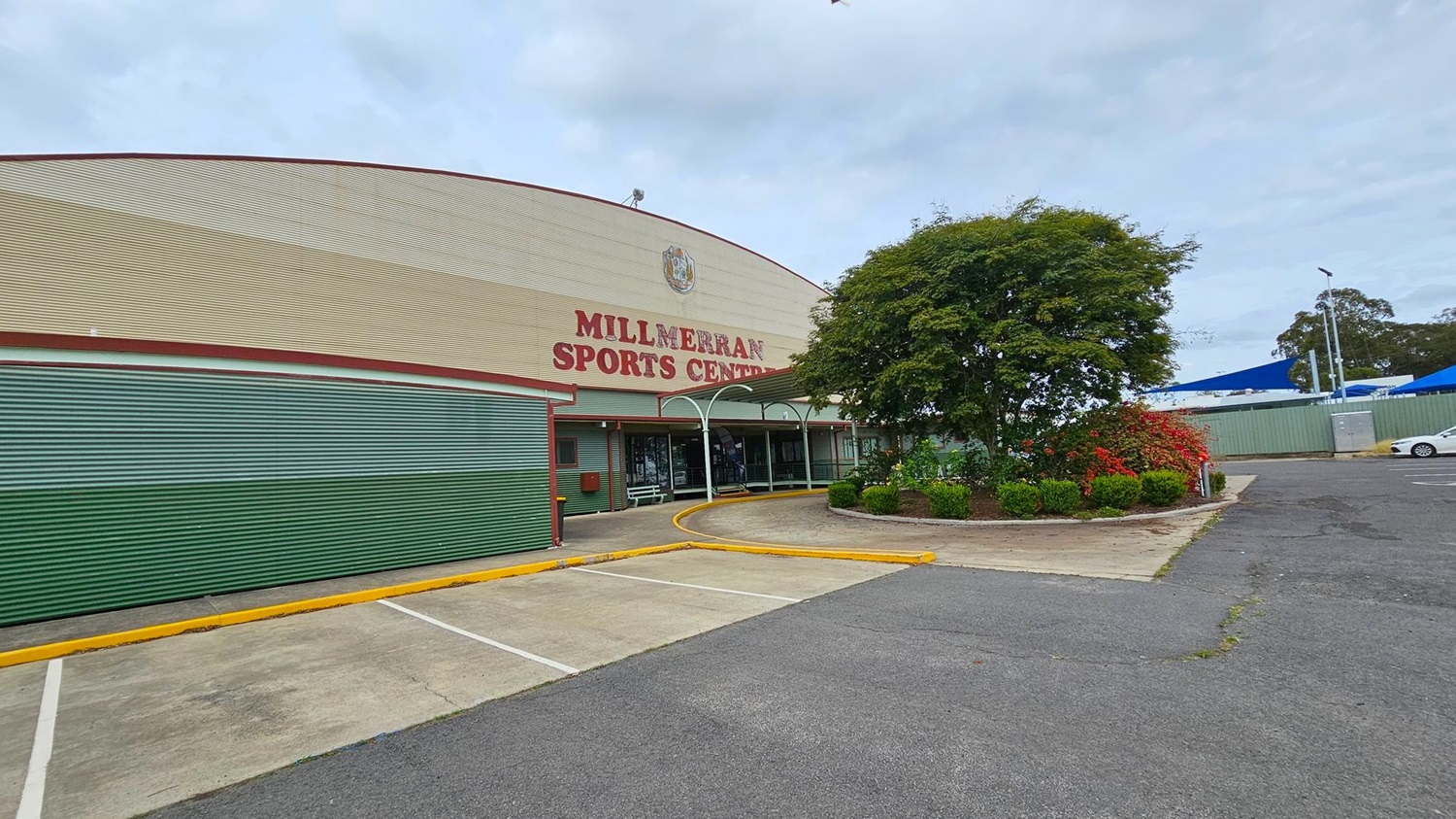 A large sports centre building is on the left, with "Millmerran Sports Centre" written on it. The building has a curved roof and green lower walls. In front, a black SUV is parked in an empty car park. On the right, a footpath runs alongside a road with a few trees and shrubs. Blue shade structures and a white car are visible in the background. The area is surrounded by greenery and utility poles.