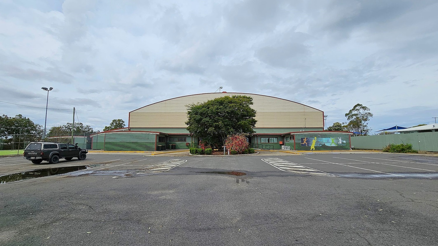A large, dome-shaped building with a green and beige exterior is in the centre. In front, a tree and some shrubs are planted in a small landscaped area. To the left, a black Isuzu vehicle is parked near a puddle on the asphalt. On the right, a mural with colourful figures is painted on the building's wall. The surrounding area is a mostly empty car park with marked spaces. Trees and a fence are visible in the background.