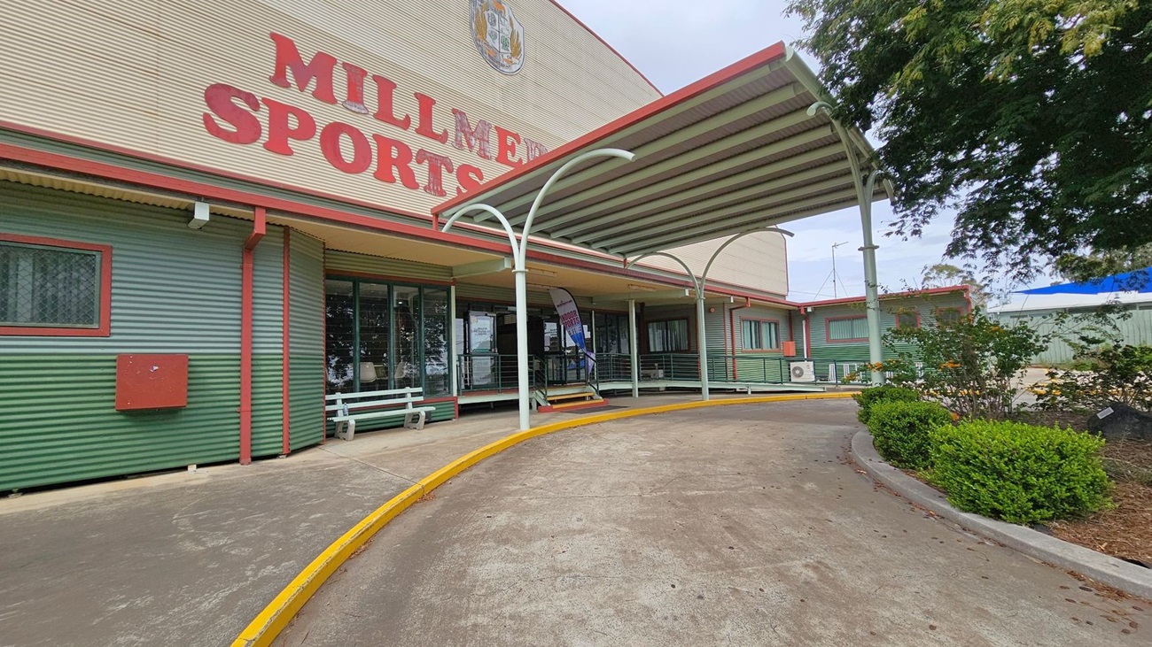 A sports building with a corrugated metal exterior painted in green and red. The large red letters on the building read "MILLMERRAN SPORTS". A covered walkway with a metal roof extends from the entrance, which has glass doors and windows. A curved concrete path with a yellow edge leads to the entrance. On the right, there are bushes and small trees. The building has a flat roof, and an air conditioning unit is visible on the wall.