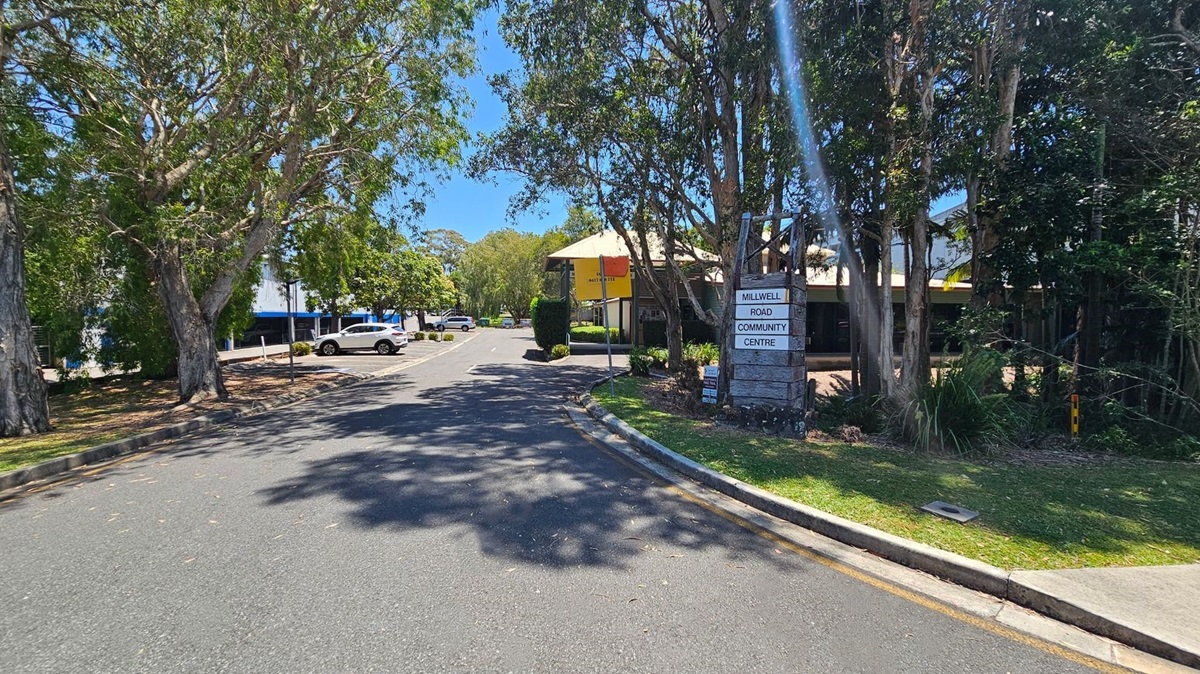 A tree-lined road with a sign on the right reading "Millwell Road Community Centre". The road curves slightly, with parked cars visible in the distance on the left. The area is shaded by large trees, and a building with a yellow awning is partially visible on the right. The ground is covered with scattered leaves, and the sky is clear.