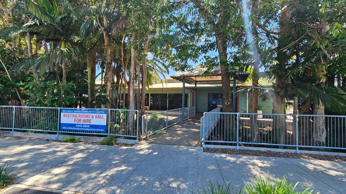 A community centre is partially obscured by tall trees and lush greenery. In the foreground, a concrete path leads to a ramp with metal railings, guiding towards the building's entrance. A sign on the left reads "Millwell Road Community Centre Meeting Rooms & Hall for Hire" with contact details. The building has a light green exterior with a covered entrance area. Shadows from the trees are cast on the path.