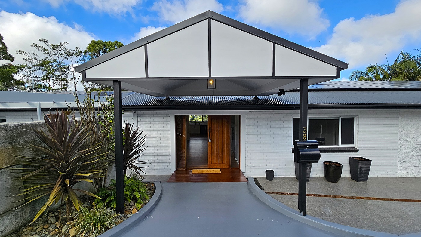On the left, there is a garden bed with various plants, including tall, spiky foliage, bordered by a concrete wall. In the middle, a grey concrete ramp leads to a wooden door set in a white brick wall. On the right, there is a black mailbox mounted on a post, a window with a black frame, and three large black plant pots. The floor is a smooth grey concrete surface, and the ceiling is a white gabled roof supported by black metal posts. The background features trees and greenery.
