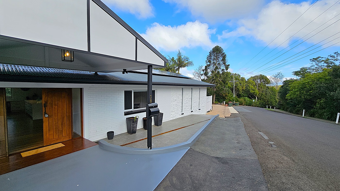 On the left, there is a wooden door with a small mat in front, leading into a white brick building. In the middle, a grey concrete ramp curves gently upwards towards the door, flanked by black plant pots. On the right, a paved road runs alongside the building, bordered by greenery and trees. The floor is a mix of grey concrete and wooden decking. The ceiling is a white gabled roof with black metal supports and a hanging light fixture. The background features tall trees and power lines.