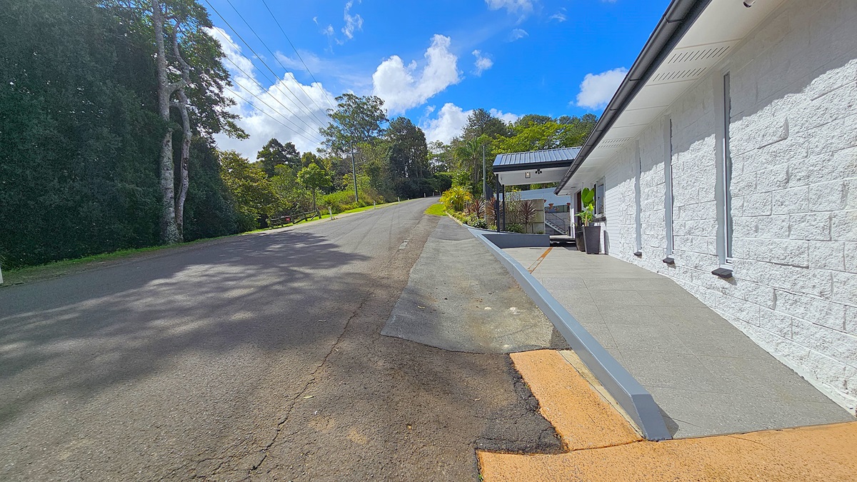 On the left, there is a paved road bordered by dense greenery and tall trees. In the middle, a sloped asphalt driveway leads to a white building with a grey roof. On the right, the building has a white brick wall with narrow windows and a grey tiled walkway. The floor is a mix of asphalt and grey tiles. The ceiling is a white overhang with ventilation slots. The background features more trees and a grassy area.