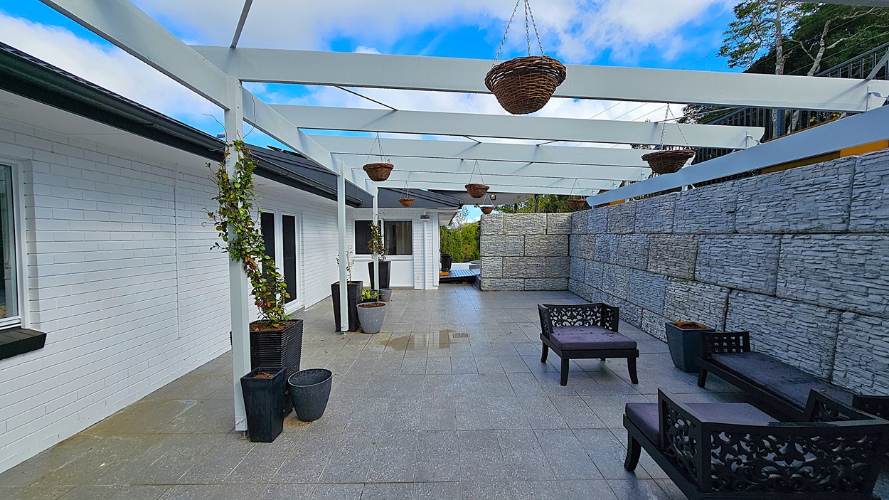 On the left, there is a white brick wall with black-framed windows and potted plants placed along the wall. In the middle, a tiled floor extends towards a white door, with hanging baskets suspended from a white pergola above. On the right, a grey stone wall runs parallel to the building, with black outdoor furniture including chairs and a table. The floor is grey tiles, and the ceiling is an open white pergola structure. The background features trees and greenery.