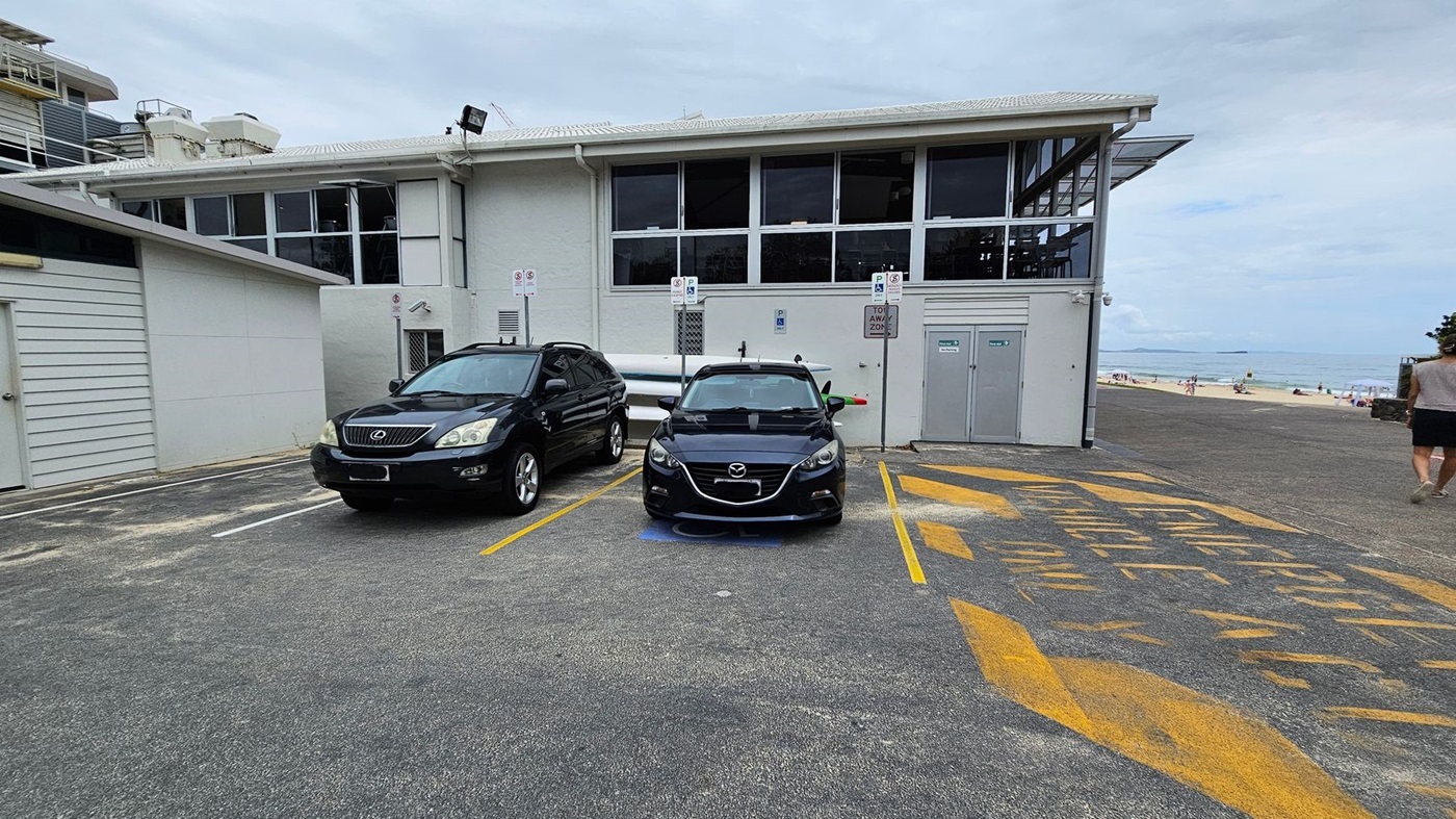 Two cars are parked in designated spaces in front of a building with large windows. The car on the left is a black Lexus, and the one on the right is a black Mazda. Yellow markings on the ground indicate a no-parking zone. To the right, a person walks towards a sandy beach with people and umbrellas visible in the distance. The building has a flat roof and light grey walls.