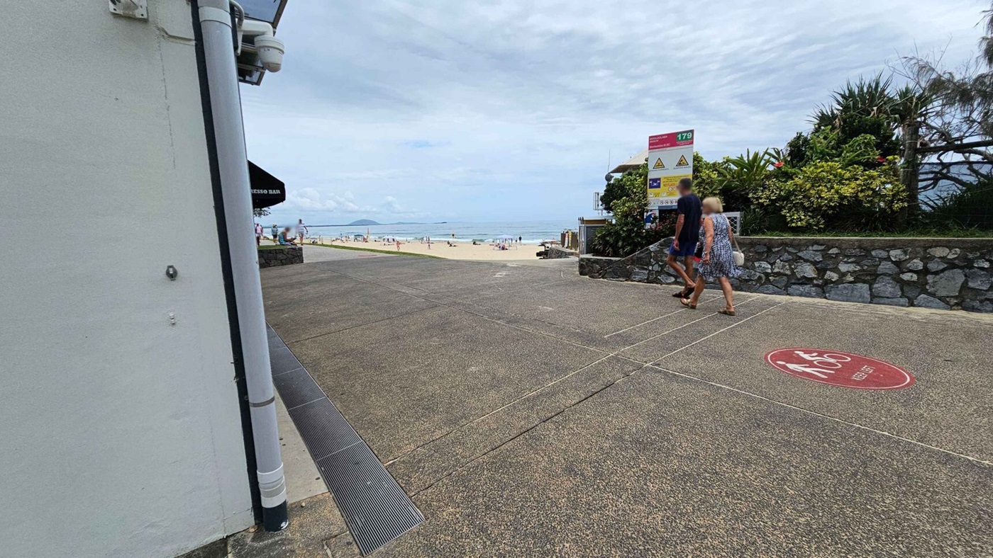 A beach scene with a pathway leading to the sand. On the left, there's a building with a pipe and a sign for an espresso bar. On the right, a stone wall with lush greenery and a sign with safety information. Two people walk along the path, which has a textured surface and a red circle with "Keep Left" written on it. In the background, people are on the beach near the ocean.