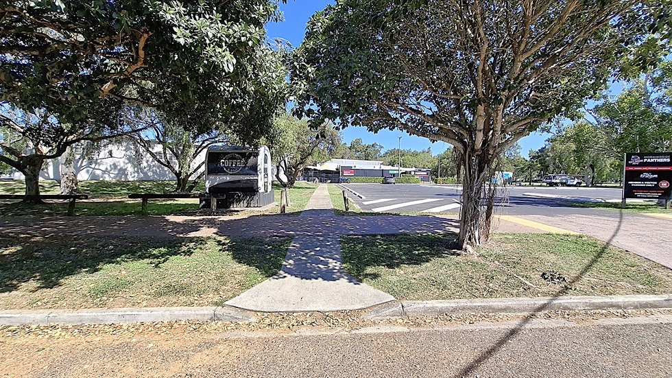 On the left, there are several large trees providing shade, with a grassy area visible behind them. A pathway leads through the greenery. To the right, a building with a white exterior is partially visible, along with a sign indicating "Panthers" and other businesses. The ground is a mix of asphalt and concrete, with a yellow tactile paving strip at the crossing.