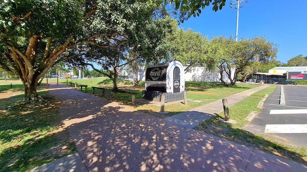 On the left, a black and white coffee truck is parked beside a path, surrounded by trees and grass. The path is made of compacted earth and is lined with benches. To the right, a concrete walkway leads towards a building with a white exterior, and further back, there is a parking area with marked spaces. The ground is a mix of grass and concrete, and the area appears well-maintained, with no visible litter.