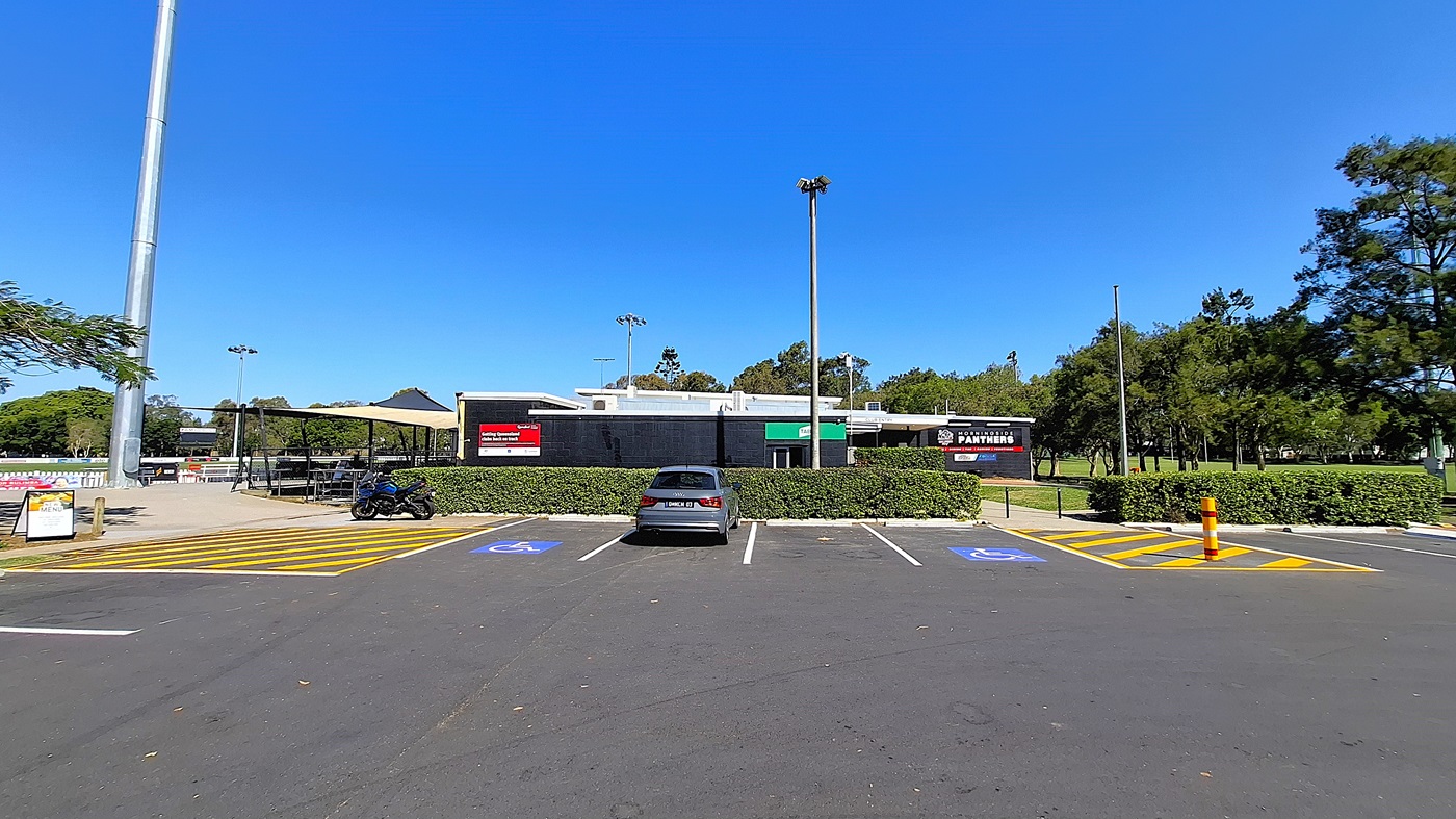 On the left, there is a parking area with a few marked spaces, including two designated for disabled parking, and a sign indicating the parking rules. A black motorcycle is parked nearby. To the right, a building with a black exterior features signs for "Panthers" and other businesses. The ground is asphalt, clearly marked with yellow lines. In the background, trees provide greenery, and there are light poles visible.