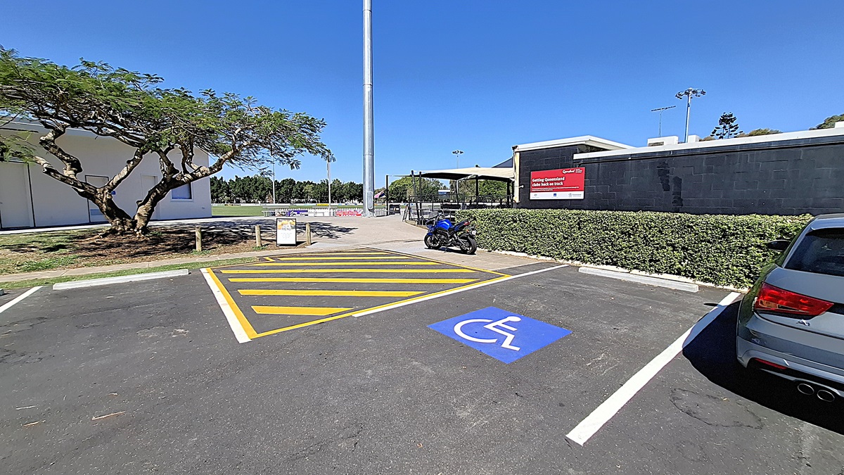 On the left, a large tree provides shade over a grassy area, with a few wooden posts visible nearby. A parking space is marked with yellow lines, including a blue symbol for disabled parking. A blue motorcycle is parked in this space. To the right, a building with a black exterior features a sign indicating "Athletics Queensland." The ground is asphalt, clearly marked, and the area appears tidy, with a low hedge bordering the parking lot.