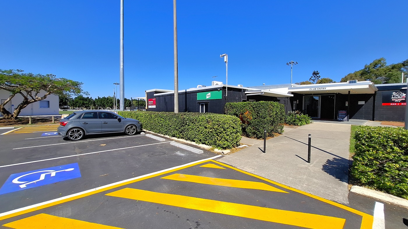 On the left, a grey car is parked in a designated disabled parking space, marked with a blue symbol. A tree with green foliage is visible behind the car, providing some shade. To the right, a building with a black exterior features a green sign indicating "Club Entry." The entrance is flanked by neatly trimmed hedges. The ground is asphalt with yellow diagonal lines marking the parking area.