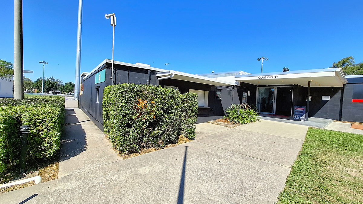 On the left, a pathway bordered by neatly trimmed hedges leads towards a building. The path is made of concrete and is well-maintained. To the right, the building features a black exterior with a sign reading "CLUB ENTRY" above the entrance. The entrance is framed by a small overhang, and there are potted plants beside the door. The ground is also concrete, with a slight incline towards the entrance.