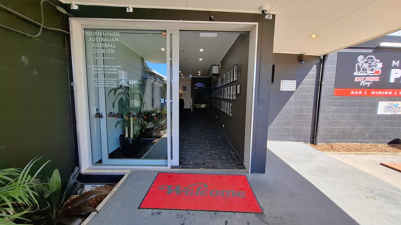 On the left, a glass door leads into a corridor with a tiled floor, and a few potted plants are positioned near the entrance. A red welcome mat is placed at the threshold. To the right, a wall displays several framed photographs, and a small air conditioning unit is mounted above. The ceiling features recessed lighting, providing illumination to the area. The floor is dark tiled, extending into the corridor, which appears clean and well-maintained.