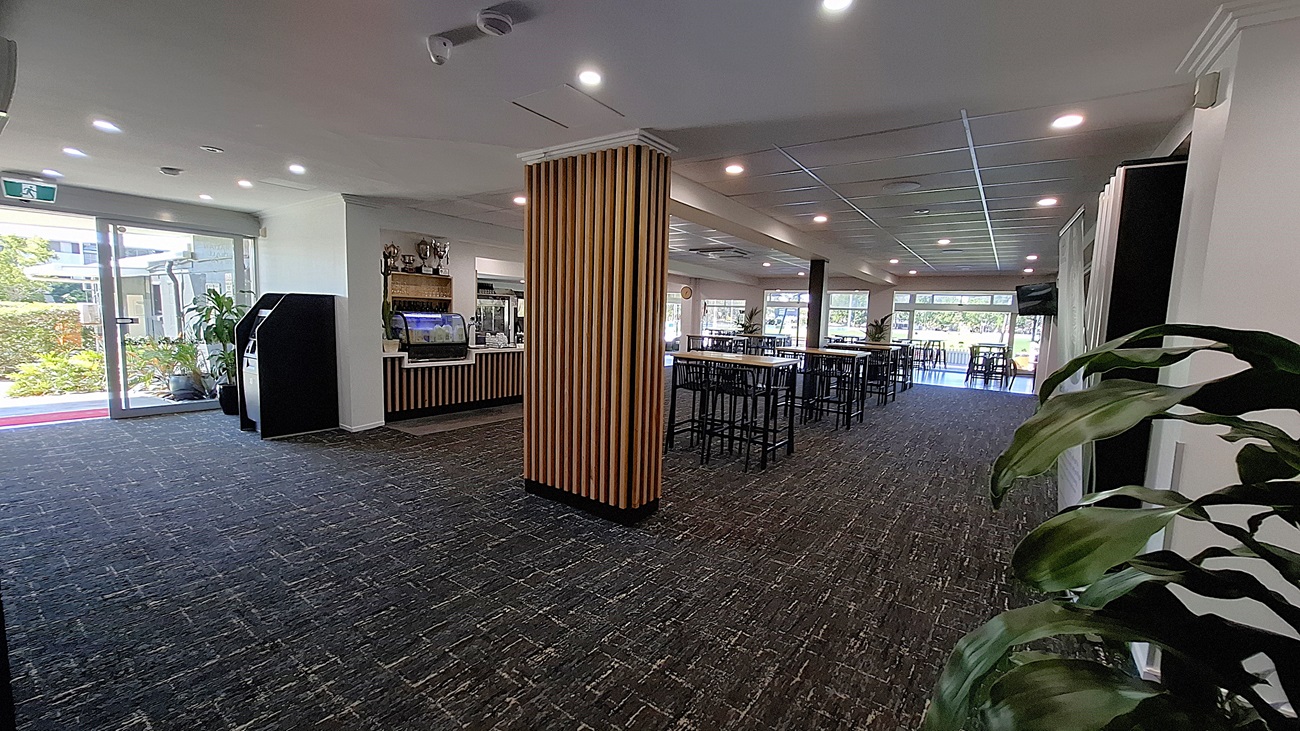 On the left, a glass door opens to the outside, with a small area featuring potted plants. Adjacent to the door, there are two black machines, possibly for ordering or payment. In the centre, a wooden bar counter with a glass display case is visible, showcasing items for sale. To the right, tall tables with black stools are arranged, and large windows allow light into the space. The floor is covered in dark carpet with a textured pattern, and the ceiling features recessed lighting.