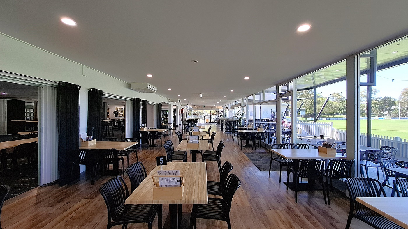 On the left, a series of black curtains frame an entrance, leading to a more private area. Several wooden tables with black chairs are arranged in the foreground, with a small menu holder on one table. In the centre, the floor is made of polished wooden boards, creating a warm appearance. To the right, large glass windows allow light to flood the space, revealing outdoor seating and greenery beyond. The ceiling is white with recessed lighting.