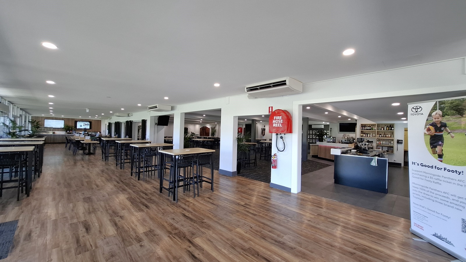 On the left, several tall tables with black stools are arranged on a polished wooden floor, creating a spacious dining area. A large window allows natural light to enter, with potted plants positioned nearby. In the centre, the floor continues with the same wooden finish, leading towards a bar area. To the right, a wall features a large banner promoting a local event, alongside a black counter with bottles displayed behind it. The ceiling is white with recessed lighting.