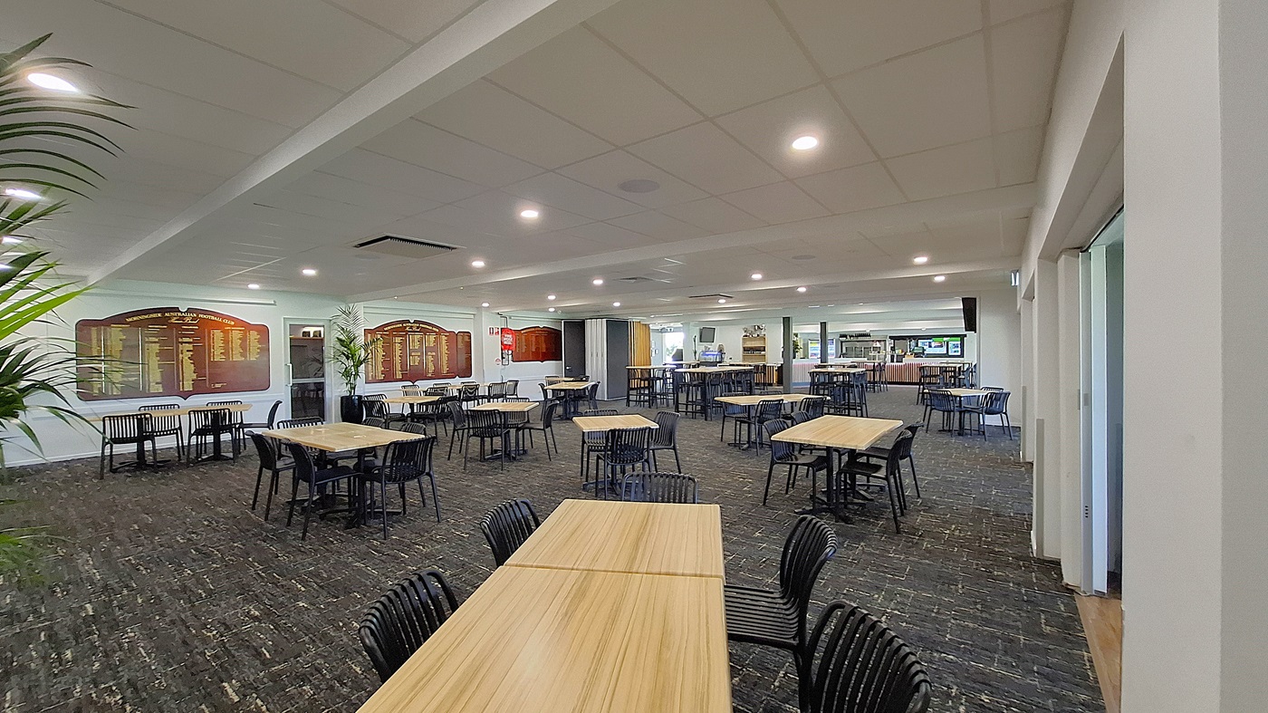 On the left, a large potted plant adds a touch of greenery beside a wall featuring framed boards with text. Several wooden tables with black chairs are arranged across the dark carpeted floor. In the centre, the space is open, with more tables visible, creating a dining area. To the right, a bar counter is seen in the background, with shelves and equipment. The ceiling is white with recessed lighting, providing a bright atmosphere throughout the room.
